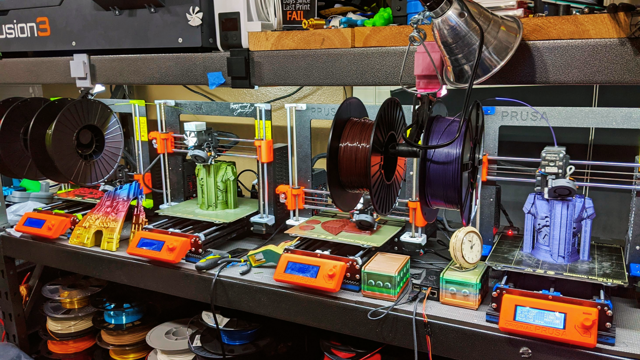 Row of four orange Prusa printers above spools of filament printing colorful 3D prints.